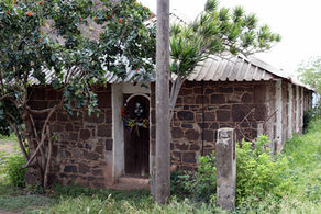 View of the narrow end of a long single-storey grey stone building, with a light grey corrogated roof. A narrow deepset doorway features a black and white image of an African person's face with a wreath of real flowers around it. The building is surrounded by lush vegetation, some of which is growing up the walls, and broken-down fence with barbed wire. In the left background is a steep hill. 