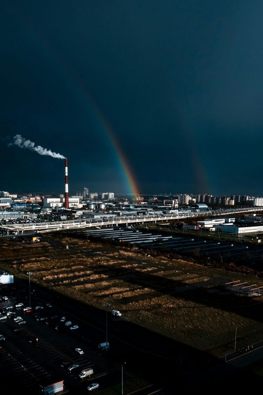 Paysage industriel sous ciel noir, fumée d’une cheminée et arc-en-ciel au-dessus de bâtiments et parkings.