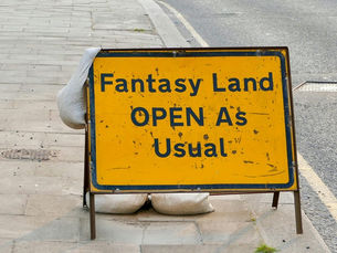 Panneau jaune sur le trottoir, texte en noir "Fantasy Land OPEN As Usual", soutenu par sacs de sable. Route grise en arrière-plan.