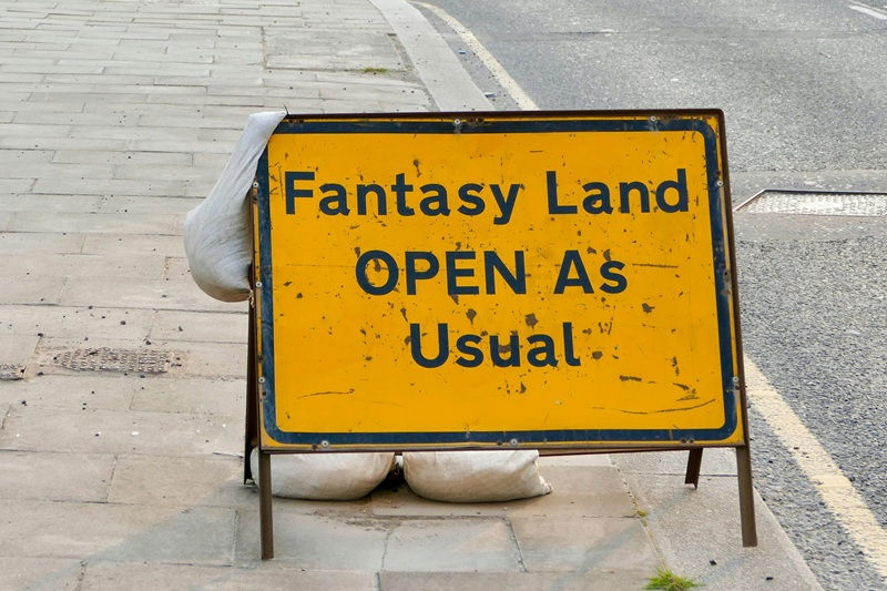 Panneau jaune sur le trottoir, texte en noir "Fantasy Land OPEN As Usual", soutenu par sacs de sable. Route grise en arrière-plan.