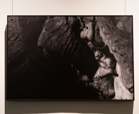 SCP - Number: Unknown by Roee Baldinger (Cerno Argus). High-contrast dark fine art photography of a small humanoid silhouette shape appearing within the lower-right quadrant of heavy, porous gray rock textures. Part of the Psychosis of a Matter sub-series.