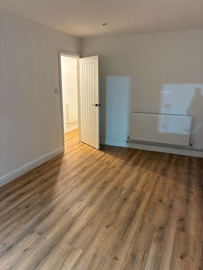 Bright newly refurbished room with warm oak-effect laminate flooring, fresh white painted walls, modern white panel door with black hardware and radiator, showcasing professional internal renovation and property refurbishment work completed by CDAA Services renovation specialists in South Wales, serving clients across the UK.