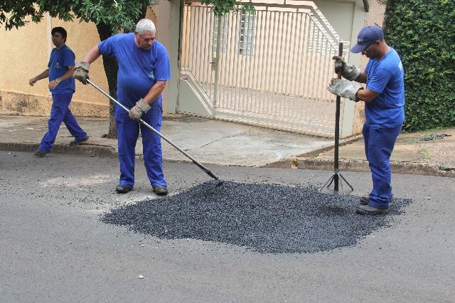 Dois funcionários da secretaria de Obras de Bauru fazendo o recapeamento de um buraco na frente de uma casa com portão branco