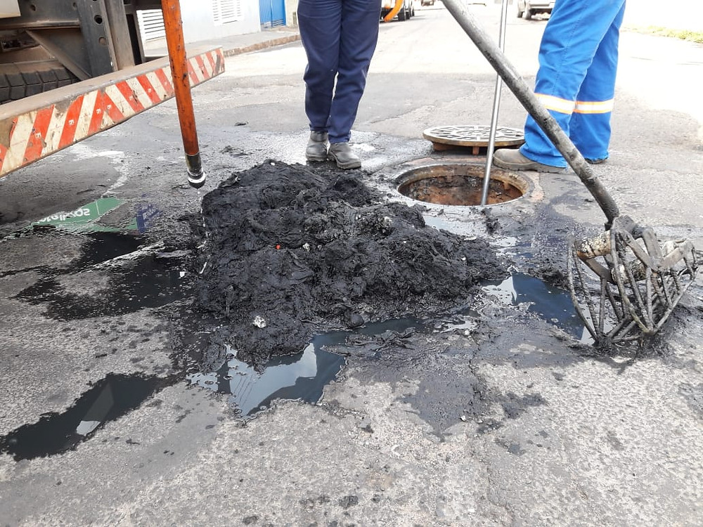 Fotografia que mostra dois profissionais trabalhando em um bueiro em uma rua de asfalto. É possível ver somente as pernas e pés dos trabalhadores que utilizam calças azuis de trabalho e botinas. Na frente deles há um bueiro aberto e ao lado está uma pilha preta de estopa retirada do bueiro. Ao lado esquerdo também é possível ver a parte de uma cancela laranja com listras brancas.
