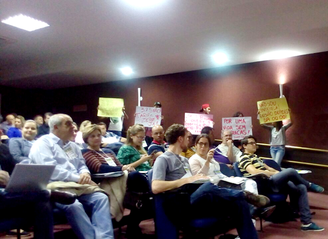 A foto mostra várias pessoas sentadas uma do lado da outra em um auditório. Ao fundo manifestantes levantam cartazes.