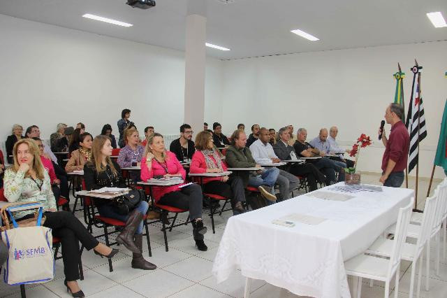 A foto mostra pessoas sentadas em fileiras horizontais. A foto é de frente. No canto direito, tem uma mesa com uma toalha branca e cadeiras brancas. Um homem branco e calvo segura o microfone. As paredes são brancas.