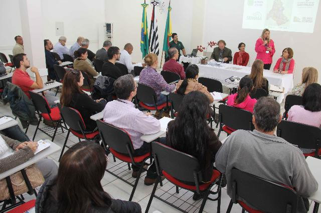 A foto mostra uma reunião em uma sala fechada. As pessoas estão sentadas em fileiras horizontais, assistindo quatro pessoas sentadas em uma mesa uma do lado da outra. Uma mulher segura o microfone e está em pé. A parede da sala é branca.