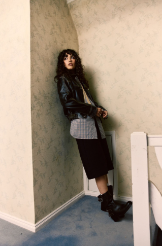 Woman in leather jacket and boots gazes upward in a narrow hallway with patterned wallpaper and blue carpet, exuding a contemplative mood.