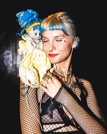 Woman with blonde and blue hair wearing fishnets holds yellow clown doll on her shoulder