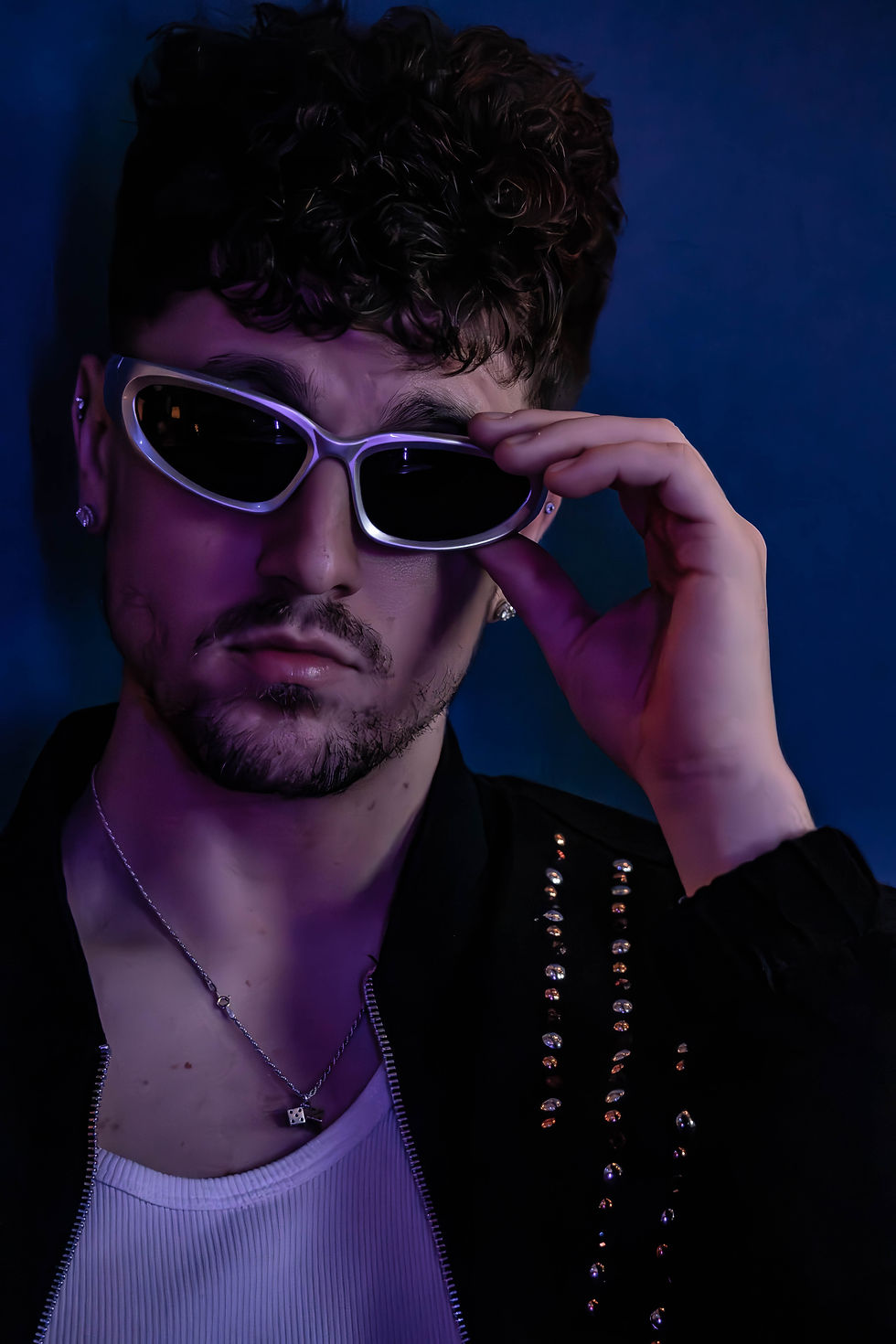Man in studded jacket adjusts white sunglasses against a dark blue background, with a purple light casting a moody glow.