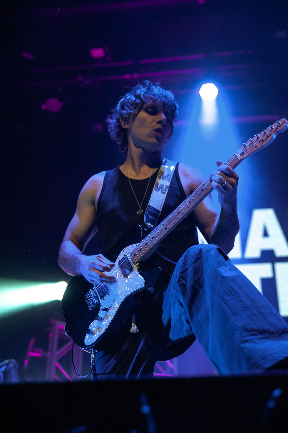 Musician playing electric guitar on stage, wearing a black tank top. Blue and purple lights create an energetic concert atmosphere.