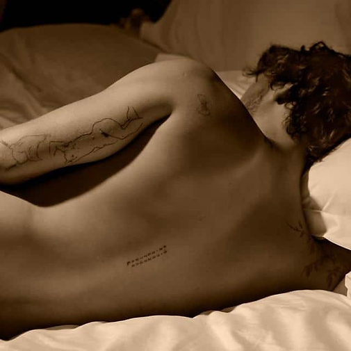Sepia-toned portrait of a man lying in bed with his back to the camera. His hair is brown and wavy, and he has a beard. He has a few tattoos on his biceps and on his spine.