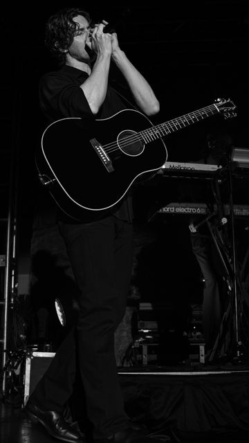Musician on stage, playing harmonica and holding a black guitar. Dark background with keyboards nearby. Intense and focused mood.