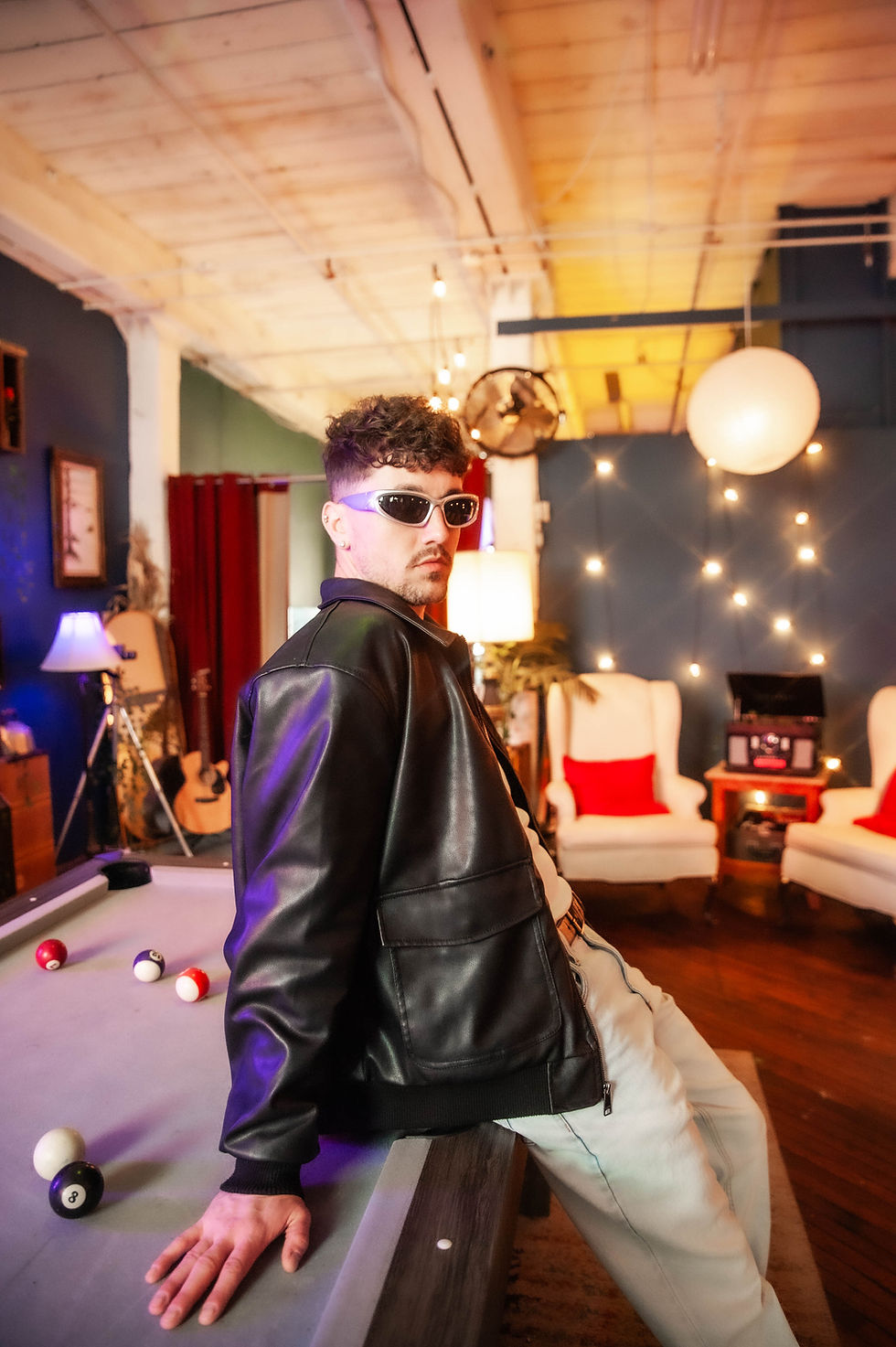 Man in sunglasses and leather jacket leaning on a billiard table. Cozy room with warm lighting, white chairs, and red pillows in the background.