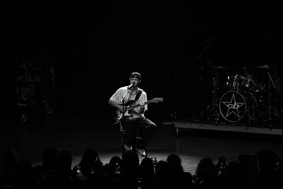 Musician playing guitar on stage with microphone, wearing a cap. Drum set with star logo in background. Audience in foreground.