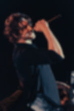 Singer with curly hair passionately performs on stage, holding a microphone. Audience captures the moment with phones. Dark, energetic atmosphere.