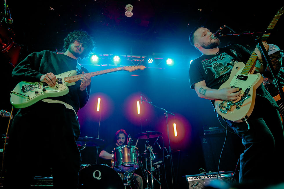 Three musicians perform on stage with electric guitars and drums. Neon lights glow blue and orange, creating a vibrant atmosphere.