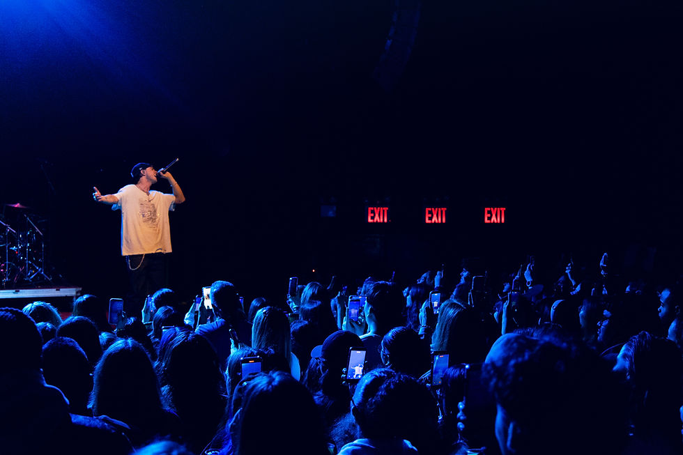 Singer in a white shirt performs on stage under blue lights, facing a lively crowd holding smartphones. Red "EXIT" signs in the background.