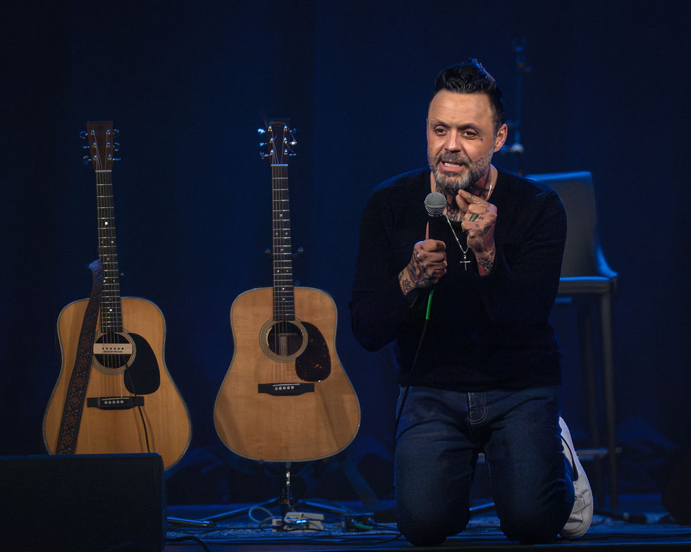 Man kneels on stage with microphone, surrounded by two acoustic guitars. Dark background, casual attire, expressive and engaged mood.