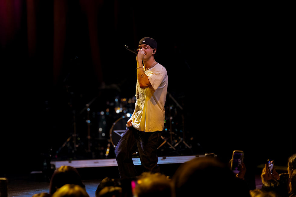 Performer on stage singing into a mic, wearing a white shirt and cap. Dimly lit background with a drum set. Audience capturing the moment.