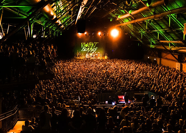 Concert scene with "Violent Femmes" on stage, large crowd, warm lighting. Audience fills an industrial venue, creating an energetic atmosphere.