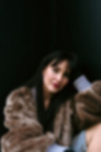 Woman with black hair in a brown fur coat poses against a dark background, looking calmly at the camera.