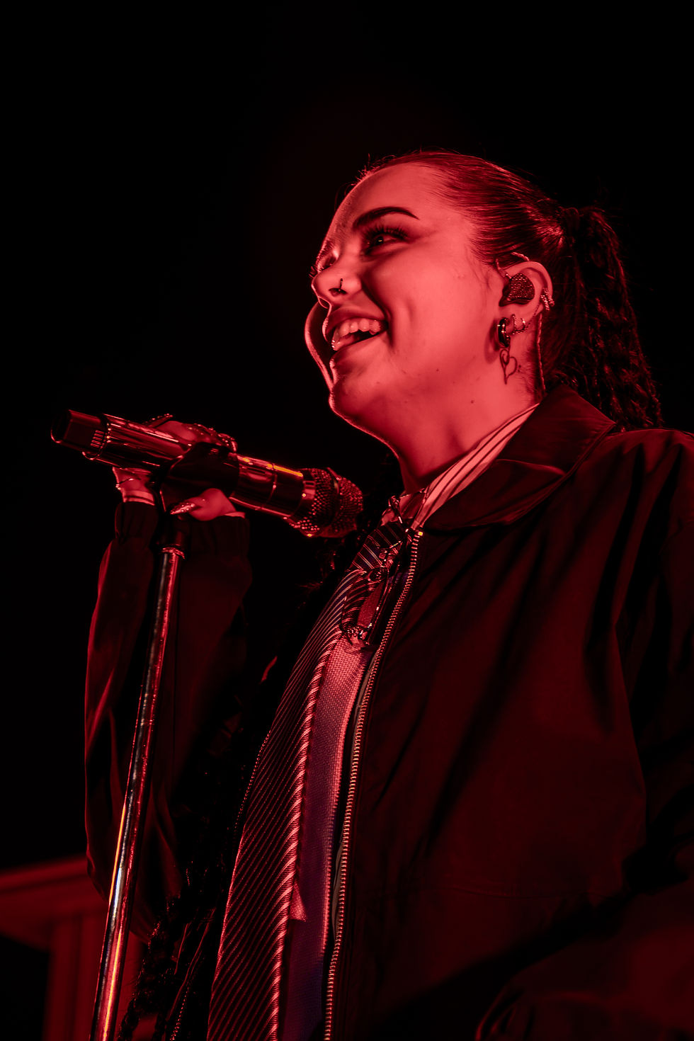 Singer smiling while performing on stage, holding a microphone. Red lighting creates an energized mood. Black background.