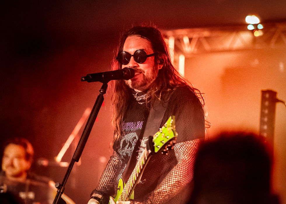 Long-haired singer in sunglasses performs into a microphone on a red-lit stage, holding a bright green guitar.