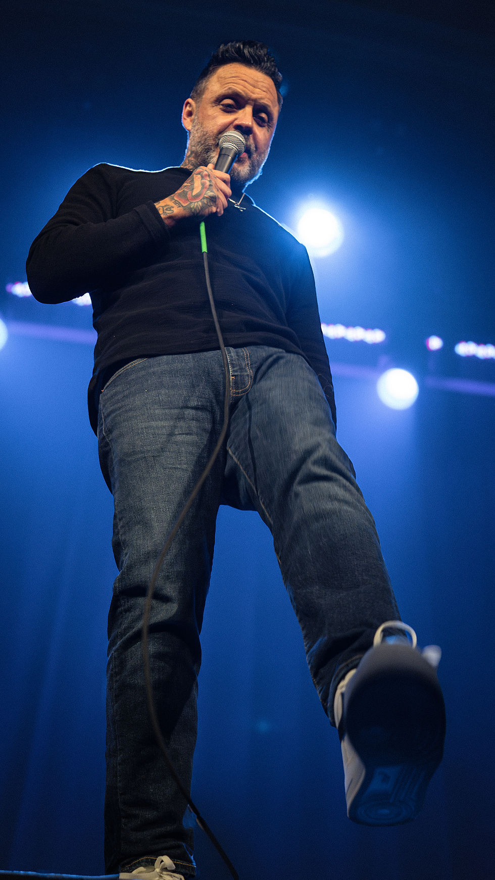 Man in black shirt and jeans sings into a mic on stage, blue lighting, dynamic low-angle shot, capturing intense performance mood.
