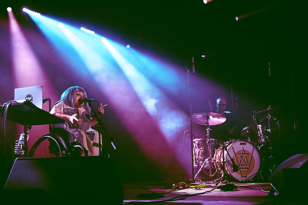Singer in a wheelchair performs onstage under purple and blue spotlights, with a drummer behind and a dark concert backdrop.