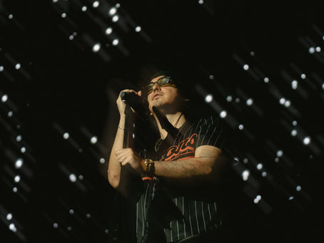 Singer in glasses grips a microphone on a dark stage, lit by scattered white lights, wearing a black pinstripe jersey with red lettering
