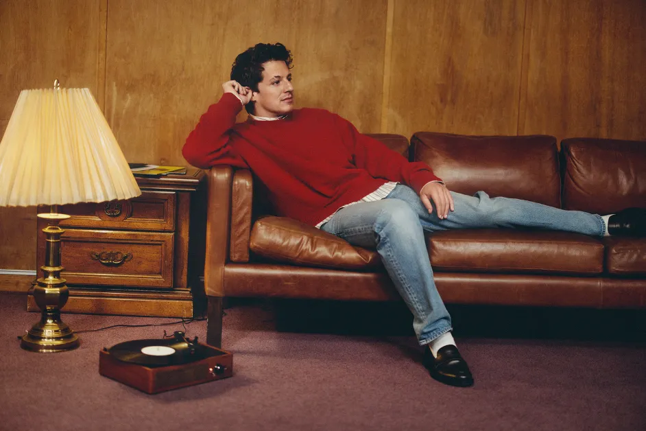 Man in red sweater lounges on brown leather sofa in a wood-paneled room. A lamp, record player, and side table add a retro vibe.