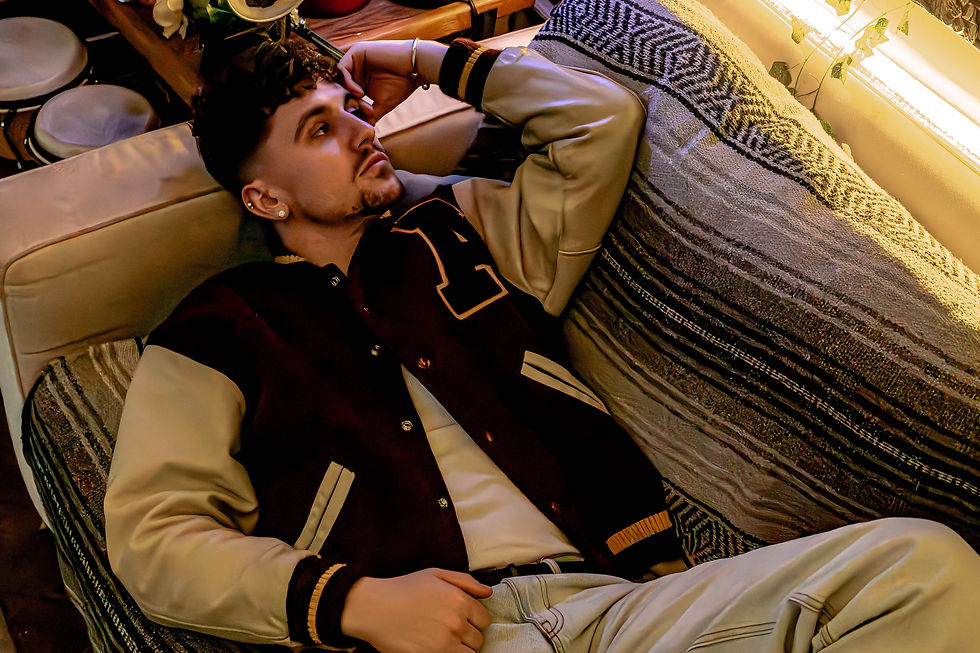 Young man in a varsity jacket relaxes on a striped couch, gazing thoughtfully. Warm lighting creates a cozy atmosphere.