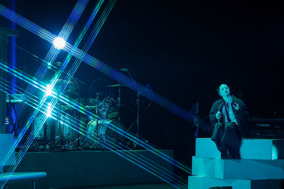 Musician on stage with a drum set and singer sitting on steps, under blue lights and beams. Calm atmosphere, visible "DiGiCo" text.