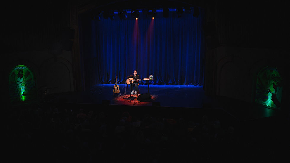 Solo performer plays guitar on a dimly lit stage with blue curtains and red spotlight. Green archway lights with artistic designs flank the sides.
