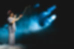 Performer in striped outfit sings on stage with dramatic blue lighting and fog, conveying a serene mood against a dark background.