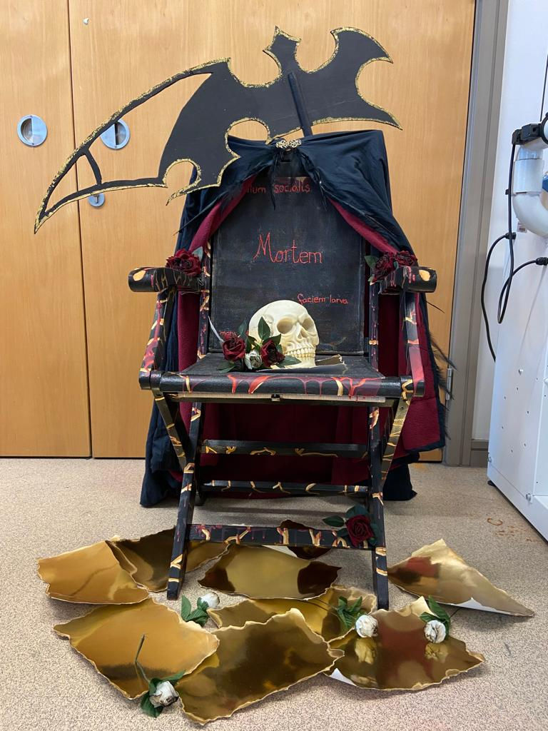 a black chair with a skull on the seat, a cloak draped over the back and a scythe standing behind it