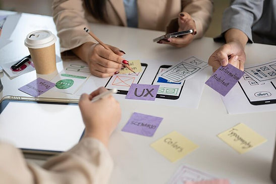 A close-up of a design team's hands working on mobile app prototypes with various notes like "UX," "Information Architect," and "Colors" on sticky notes.