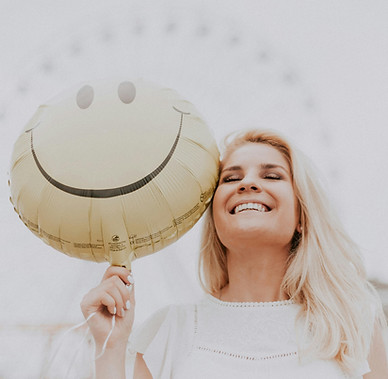 Smiling blonde woman holding a happy face balloon, symbolising emotional wellbeing and joy in mental health recovery
