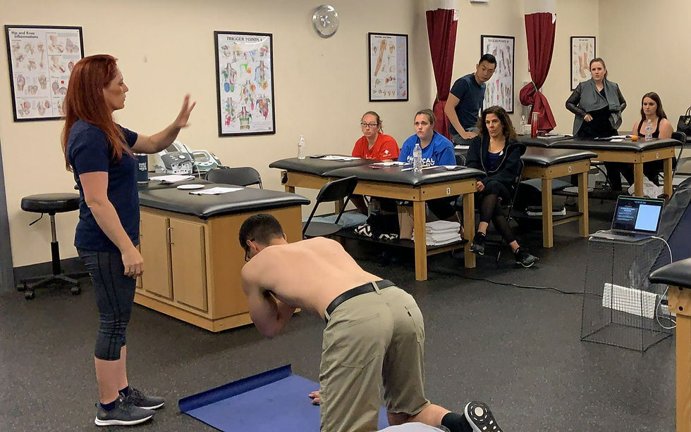 A woman instructs a seated, shirtless man in a classroom. Students watch, with anatomy charts on the walls and a laptop on the floor.