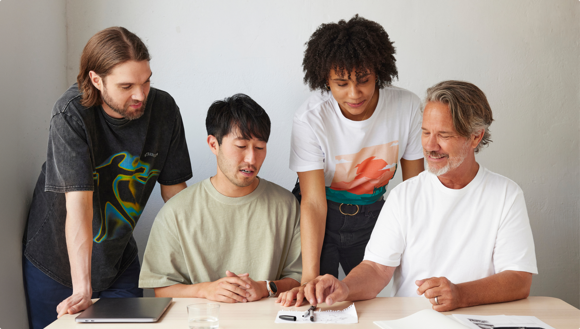 Four people are having a discussion. Two of them are sitting at a table and the other two are standing behind them, they're all looking at a document. 