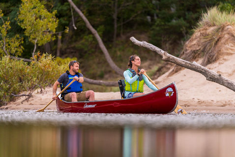 Couple paddling a red canoe along a quiet sandy shoreline.