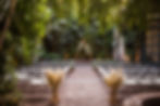 An intimate wedding ceremony setting outdoors, with a modern geometric arch and seating arranged amidst an abundance of tropical plants, creating a serene and private ambiance.