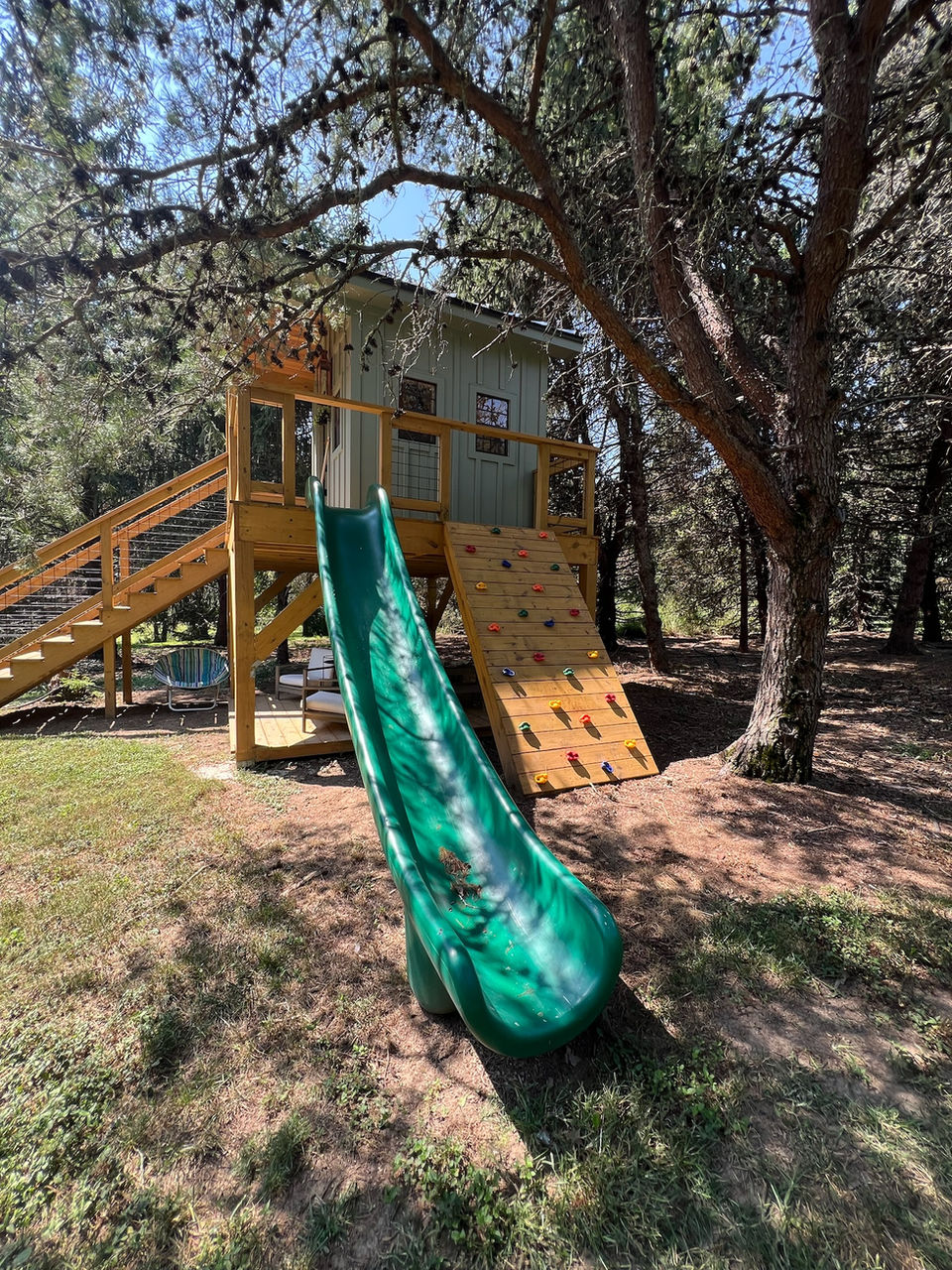 Backyard treehouse fort in Nashville with climbing wall and spiral green slide surrounded by trees.