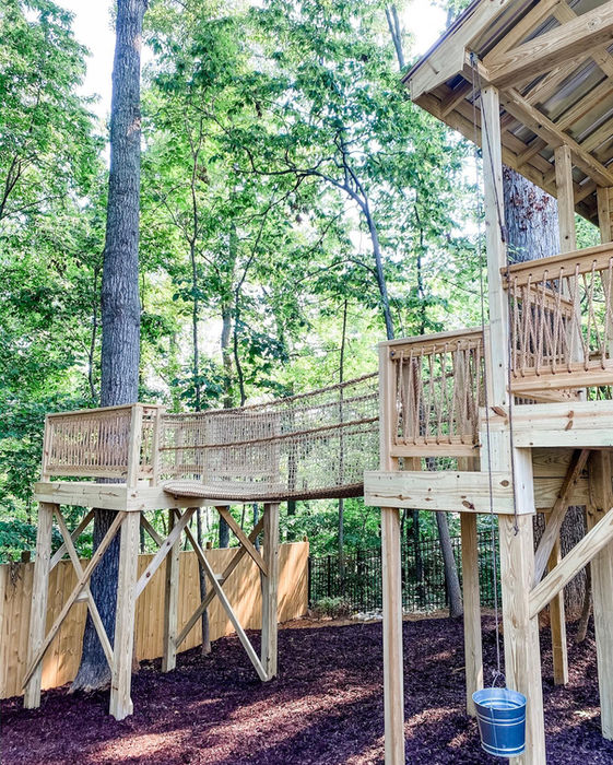 Wooden treehouse with a bridge and lush green trees in the background.