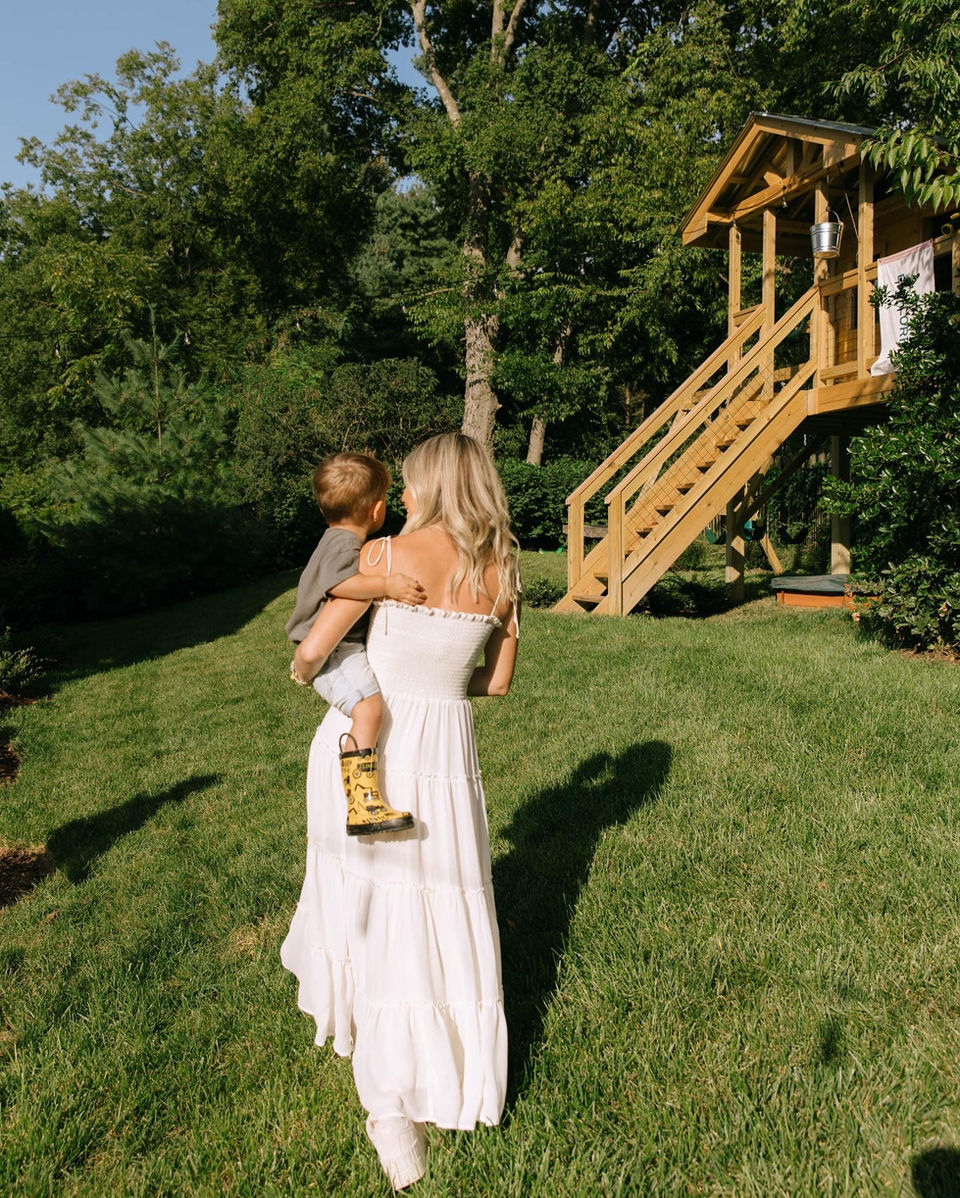 Woman carrying child, walking towards treehouse on a sunny day.