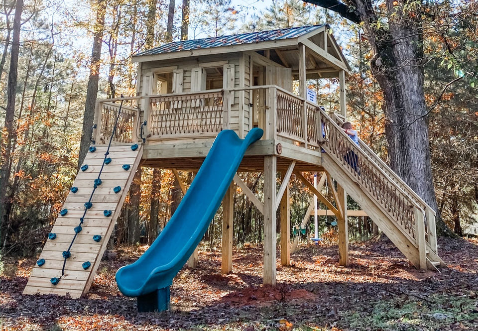 Wooden treehouse with a blue slide, climbing wall, and stairs in the woods. Firefly Forts