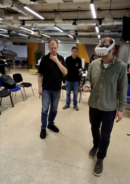 A photo from the Video Game Accessibility Symposium. On the right, a user wears a white VR Meta Quest headset and a green shirt, holding a Quest controller. On the left, a member of the Virtual Guide Dogs team in a black polo shirt watches Paul’s movements with a thoughtful expression. In the background, blue chairs and industrial lighting fill the event space.