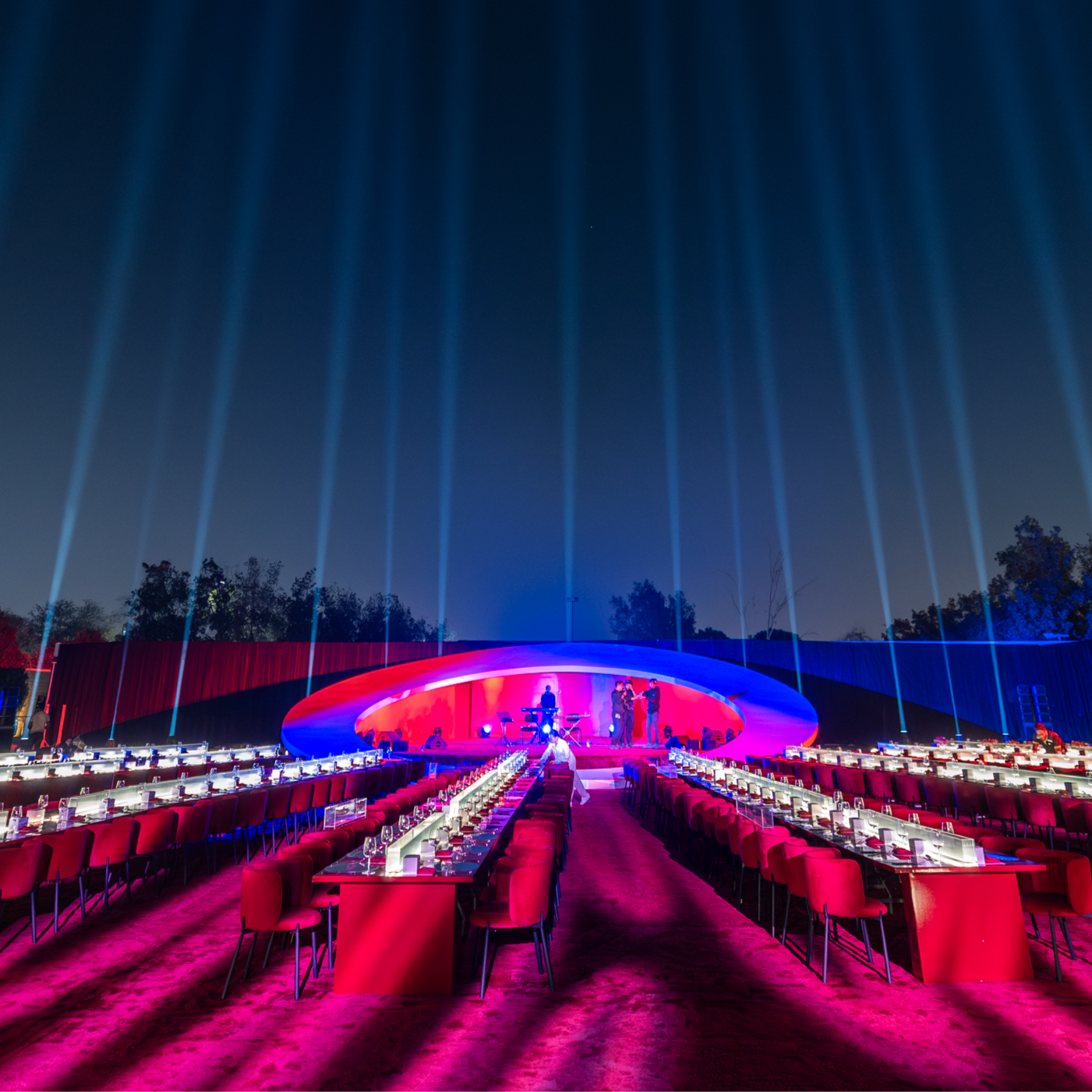 The Ring by Mishele — radial dining tables lead towards a red and blue elliptical stage beneath vertical beams of light.