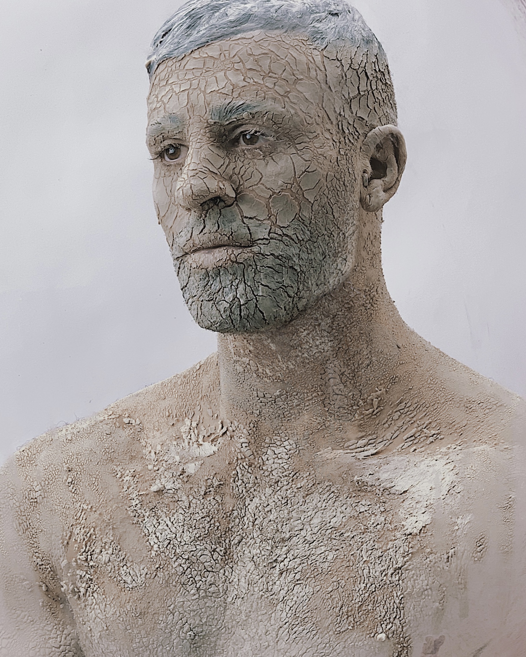 Isolation by Mishele Doueihy — portrait of the artist with his face and bare upper body covered in a cracked grey coating.